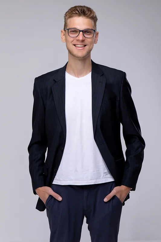 Ricardo Werlang de blazer escuro e camiseta branca, mãos nos bolsos, fundo cinza claro.