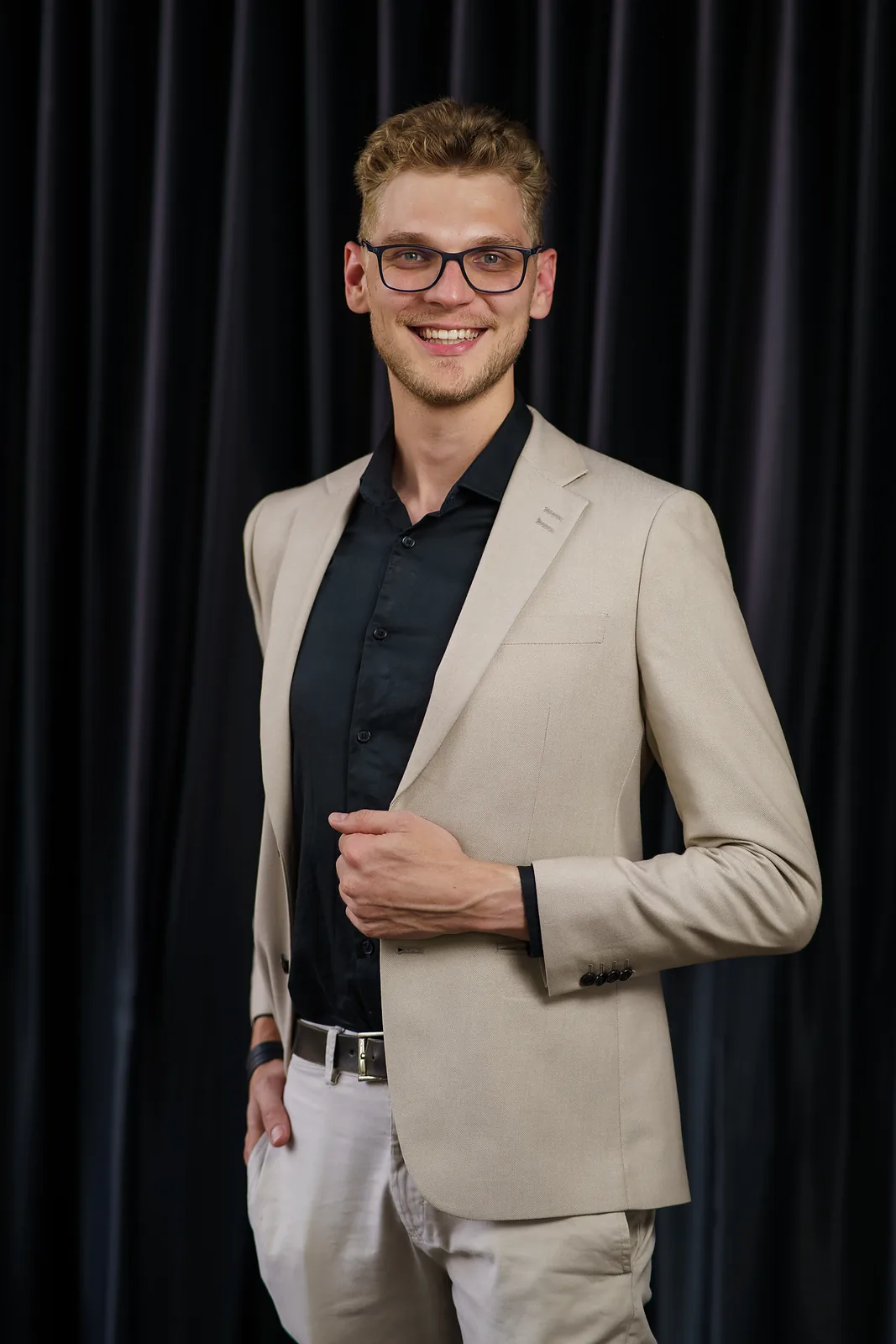 Ricardo Werlang sorrindo, blazer bege e camisa preta, cortina escura ao fundo.