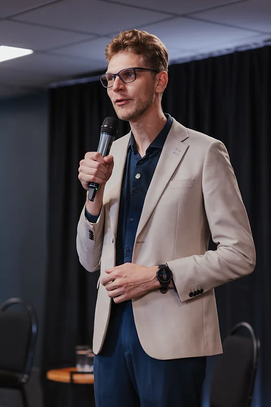 Ricardo Werlang falando ao microfone no palco, blazer bege.