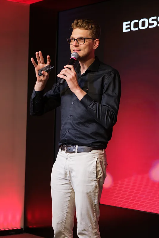 Ricardo Werlang em palestra, microfone na mão, telão vermelho.